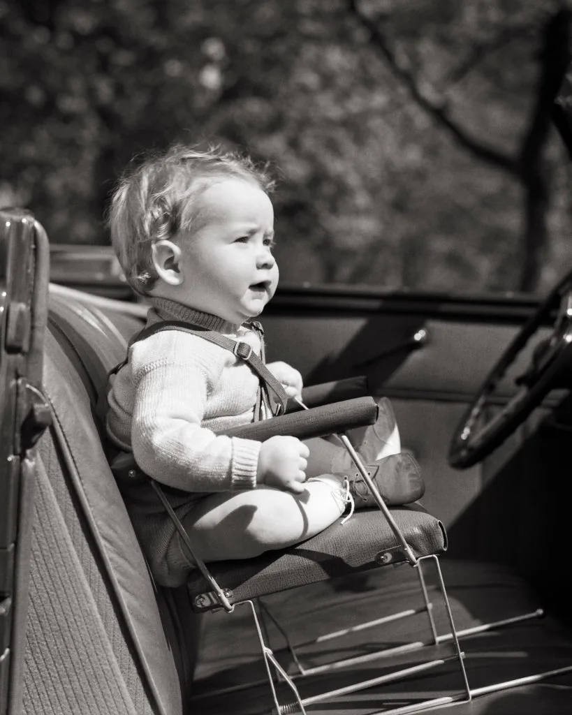 A toddler seated in a 1930s wire frame child car seat without safety restraints.