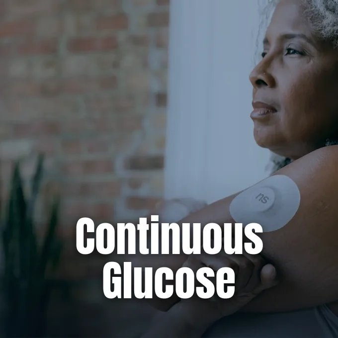 A woman wearing a continuous glucose monitor sensor on her upper arm looks thoughtfully aside.