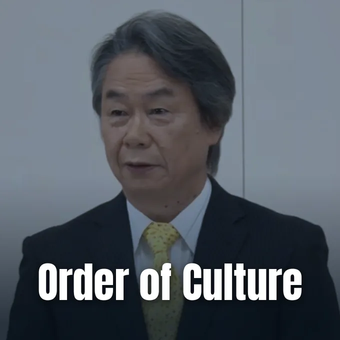 Shigeru Miyamoto wearing a formal suit and yellow tie during a press conference
