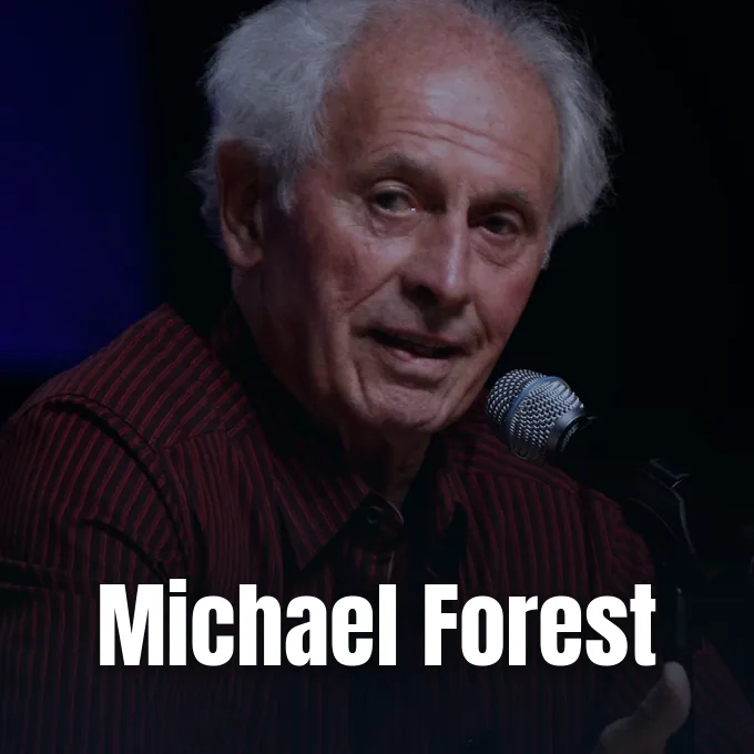 An older Michael Forest speaking into a microphone against a dark background during an appearance.