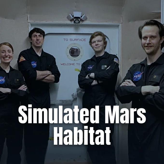 Four NASA crew members in dark uniforms standing together in front of an airlock entrance.