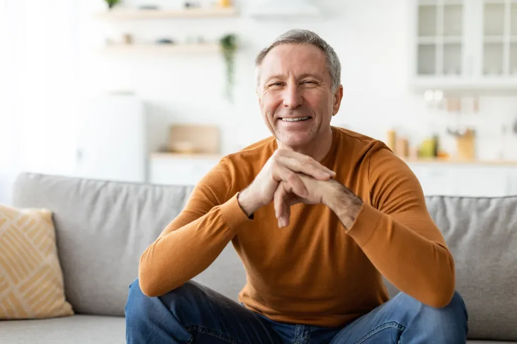 A mature man sits on a couch and smiles.