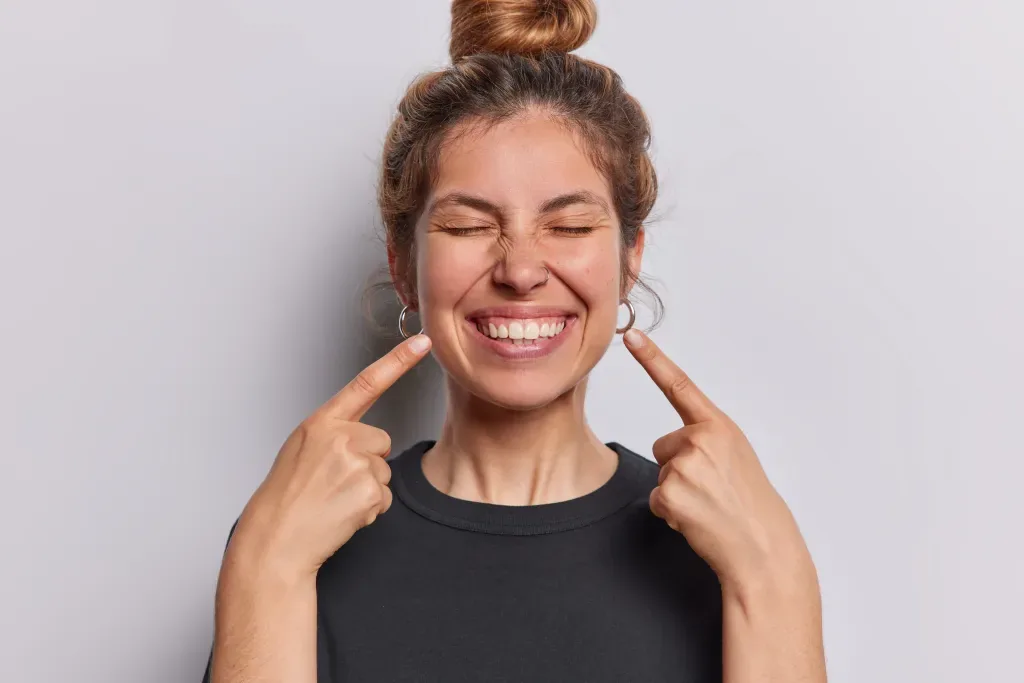 A woman gestures toward her smile to show how regular smiling influences emotional well-being.