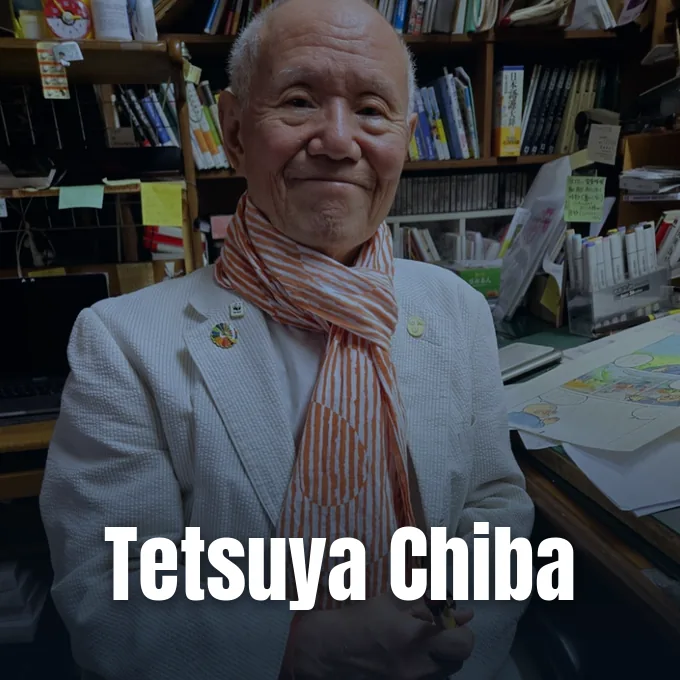 Manga artist Tetsuya Chiba smiling in his studio while wearing a scarf and blazer.